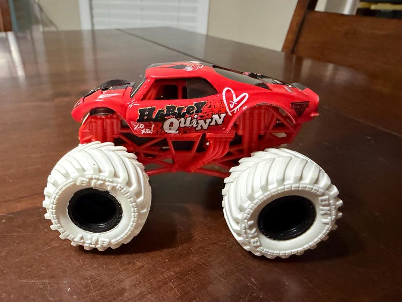 May include: A red monster truck toy with white tires. The truck features the name "Harley Quinn" in white and black lettering, along with a heart and "xo xo" accents. The tires have black centers.