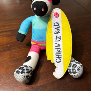 May include: A plush cow toy wearing sunglasses and a green and yellow visor, a blue and turquoise striped shirt, and pink shorts. The cow is holding a yellow surfboard with the text "CHIKIN IZ RAD". The cow's feet have a black and white checkered pattern.
