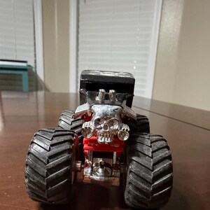 May include: A toy monster truck with large black tires and a silver skull-shaped grill. The truck has a black cab and red accents, giving it a menacing appearance. This toy is designed for play and collecting.
