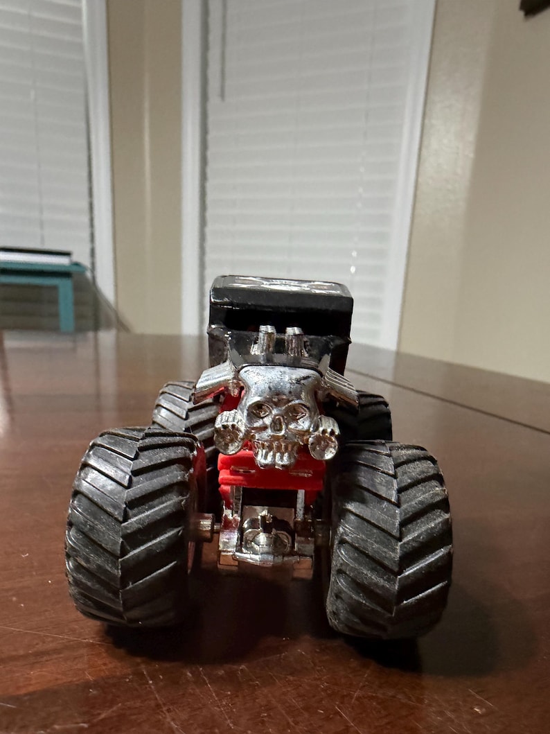 May include: A toy monster truck with large black tires and a silver skull-shaped grill. The truck has a black cab and red accents, giving it a menacing appearance. This toy is designed for play and collecting.