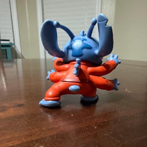 May include: A blue and red Stitch figurine, a character from the animated film Lilo & Stitch. The figurine has large blue ears, four arms, and is wearing a red jumpsuit with blue accents. The figure is posed on a dark wooden surface.
