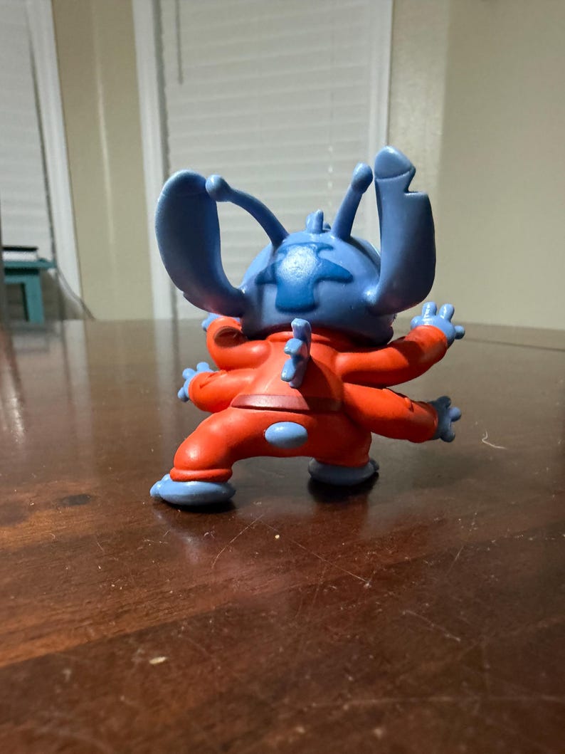 May include: A blue and red Stitch figurine, a character from the animated film Lilo & Stitch. The figurine has large blue ears, four arms, and is wearing a red jumpsuit with blue accents. The figure is posed on a dark wooden surface.