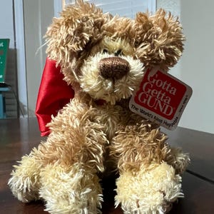 May include: A plush, seated dog toy with shaggy, light brown fur and a red ribbon. The toy has a brown nose and a tag that reads "Gotta Getta GUND". The toy is on a dark brown surface.