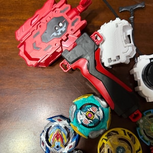 May include: A collection of colorful spinning top toys and launchers. The tops feature various designs with blue, yellow, green, and red accents. Launchers are red, black, and white. The toys are designed for competitive play.