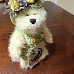 Pode incluir: Um urso de pelúcia creme usando um chapéu e vestido florais. O chapéu apresenta flores amarelas, azuis e creme. O vestido é verde com um padrão floral. Uma etiqueta com as palavras "Boyds Bears Bearwear" está anexada.