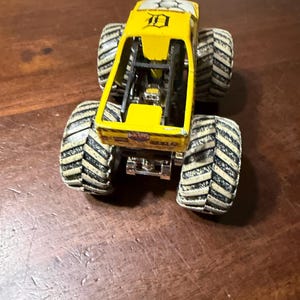 Puede incluir: Un cami&oacute;n monstruo de juguete amarillo con neum&aacute;ticos grandes en blanco y negro. El cami&oacute;n tiene una jaula antivuelco y el n&uacute;mero "10" es visible en la parte superior. El cami&oacute;n est&aacute; sobre una superficie de madera.