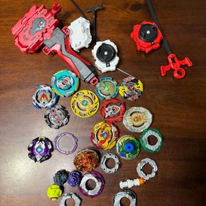 May include: A collection of colorful spinning tops and launchers in various designs. The tops feature a variety of colors including red, blue, yellow, and green. The launchers are red, white, and black.