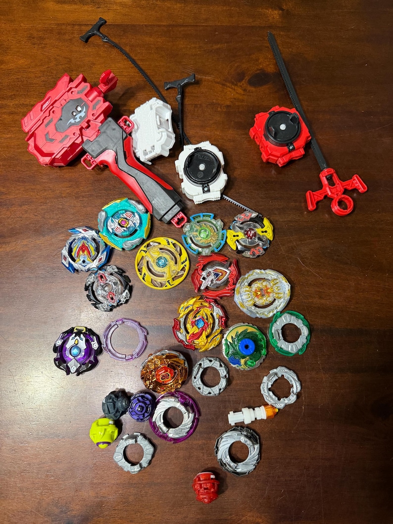 May include: A collection of colorful spinning tops and launchers in various designs. The tops feature a variety of colors including red, blue, yellow, and green. The launchers are red, white, and black.