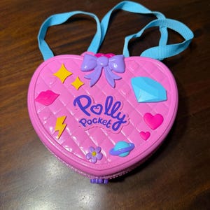 May include: A pink heart-shaped Polly Pocket case with a blue handle. The case has a quilted pattern and is decorated with a purple bow, a diamond, stars, a lightning bolt, a flower, a planet, a heart, and the words "Polly Pocket".