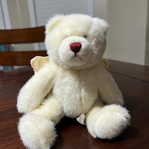 Puede incluir: Un oso de peluche color crema con una nariz roja y peque&ntilde;as alas de color amarillo claro. El peluche est&aacute; sobre una superficie de madera marr&oacute;n oscuro. El oso tiene una textura suave y esponjosa.