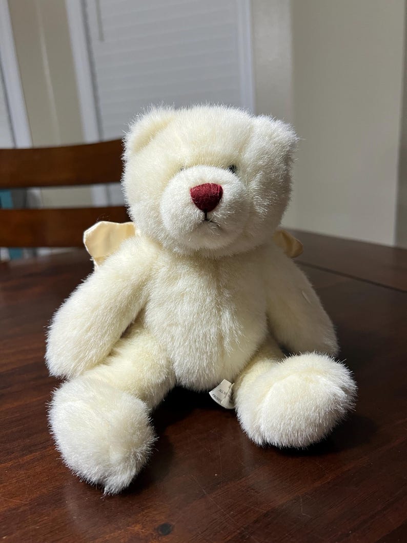 Puede incluir: Un oso de peluche color crema con una nariz roja y peque&ntilde;as alas de color amarillo claro. El peluche est&aacute; sobre una superficie de madera marr&oacute;n oscuro. El oso tiene una textura suave y esponjosa.