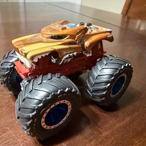 May include: A toy monster truck with a brown and yellow body, red chassis, and large black tyres. The truck features a detailed design with a front resembling a roaring animal. The tyres have blue accents.