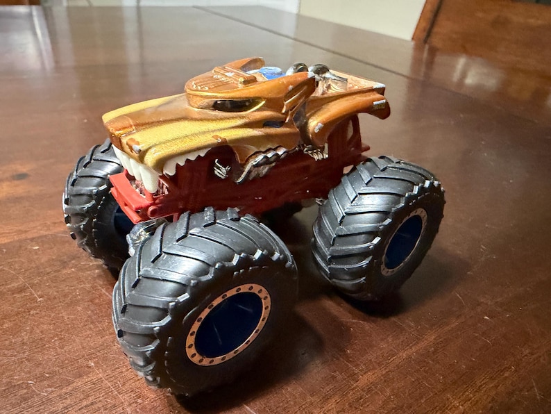 May include: A toy monster truck with a brown and yellow body, red chassis, and large black tyres. The truck features a detailed design with a front resembling a roaring animal. The tyres have blue accents.