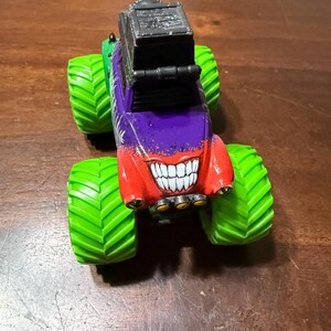 May include: A colorful toy monster truck with large green tires. The truck's body is painted in red, purple, and green, featuring a grinning mouth design. A black rectangular structure sits atop the truck.