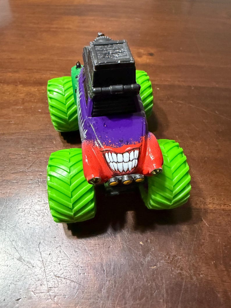 May include: A colorful toy monster truck with large green tires. The truck's body is painted in red, purple, and green, featuring a grinning mouth design. A black rectangular structure sits atop the truck.