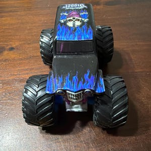 Puede incluir: Cami&oacute;n monstruo de juguete negro con neum&aacute;ticos grandes y negros. El cami&oacute;n presenta un gr&aacute;fico de calavera con un sombrero morado y llamas azules en el cap&oacute;. La palabra "Digger" est&aacute; impresa en la parte superior del cami&oacute;n.