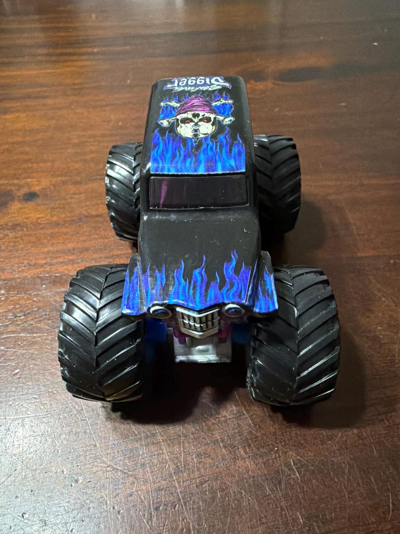 Puede incluir: Cami&oacute;n monstruo de juguete negro con neum&aacute;ticos grandes y negros. El cami&oacute;n presenta un gr&aacute;fico de calavera con un sombrero morado y llamas azules en el cap&oacute;. La palabra "Digger" est&aacute; impresa en la parte superior del cami&oacute;n.