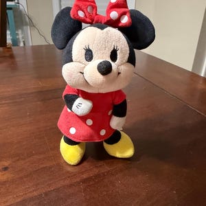 K&ouml;nnte beinhalten: Eine Pl&uuml;schpuppe von Minnie Mouse, mit einem roten Kleid mit wei&szlig;en Punkten, einer passenden Schleife und gelben Schuhen. Die Puppe hat schwarze Ohren und ein l&auml;chelndes Gesicht.