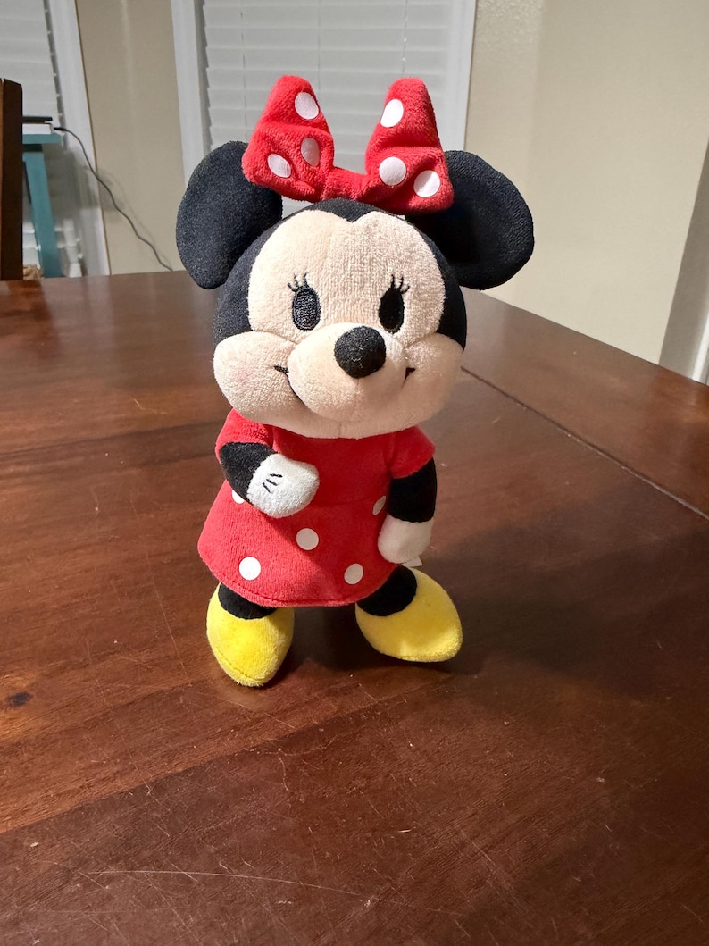 K&ouml;nnte beinhalten: Eine Pl&uuml;schpuppe von Minnie Mouse, mit einem roten Kleid mit wei&szlig;en Punkten, einer passenden Schleife und gelben Schuhen. Die Puppe hat schwarze Ohren und ein l&auml;chelndes Gesicht.
