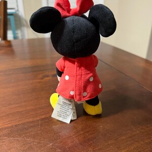 K&ouml;nnte beinhalten: Eine Minnie Mouse Pl&uuml;schpuppe, von hinten gesehen, steht auf einer Holzoberfl&auml;che. Die Puppe hat einen schwarzen Kopf mit gro&szlig;en Ohren und einer roten Schleife. Sie tr&auml;gt ein rotes Kleid mit wei&szlig;en Punkten und gelbe Schuhe.