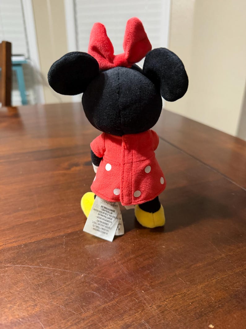 K&ouml;nnte beinhalten: Eine Minnie Mouse Pl&uuml;schpuppe, von hinten gesehen, steht auf einer Holzoberfl&auml;che. Die Puppe hat einen schwarzen Kopf mit gro&szlig;en Ohren und einer roten Schleife. Sie tr&auml;gt ein rotes Kleid mit wei&szlig;en Punkten und gelbe Schuhe.