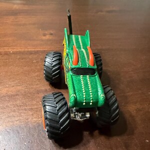 May include: Green and yellow monster truck toy with large black tires and orange accents. The truck has a detailed design with a dragon-like appearance, including horns and a textured body. The toy is on a wooden surface.