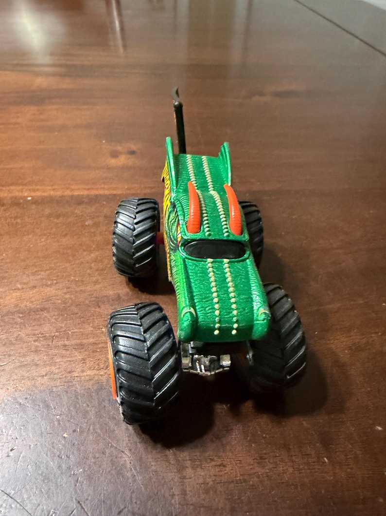 May include: Green and yellow monster truck toy with large black tires and orange accents. The truck has a detailed design with a dragon-like appearance, including horns and a textured body. The toy is on a wooden surface.