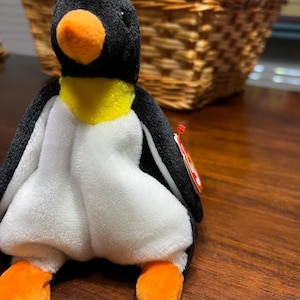 Puede incluir: Un juguete de peluche de pingüino con cabeza, espalda y alas negras, vientre blanco y pies y pico naranjas. El pingüino tiene un cuello amarillo y una etiqueta roja adjunta. El juguete está sobre una superficie de madera.