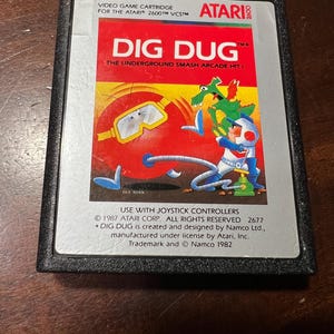 May include: A vintage Atari 2600 video game cartridge for the game "Dig Dug." The label features a colorful illustration of the game's characters and logo against a red and yellow background. The cartridge is black.