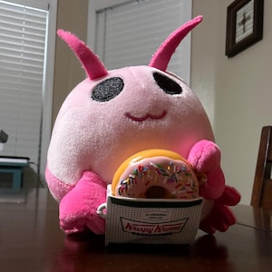 May include: A pink plush creature with large black eyes and pink antennae holds a Krispy Kreme box with a donut. The donut is pink with colorful sprinkles. The box has the words "Krispy Kreme" on it.