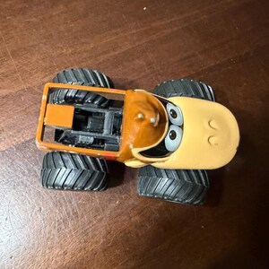 May include: A toy monster truck with a cartoon design. The truck has a tan cab with a brown top and large black tyres. The truck's face has large eyes and a smiling expression. The truck is orange and black.