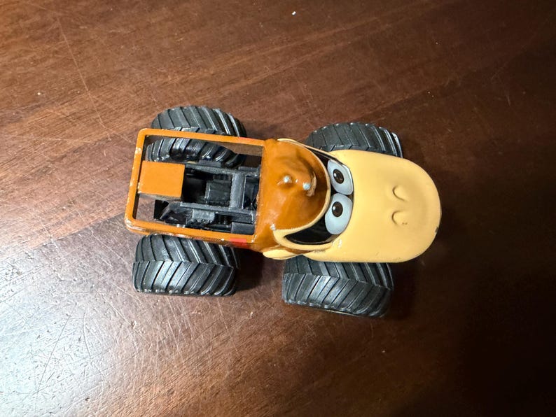 May include: A toy monster truck with a cartoon design. The truck has a tan cab with a brown top and large black tyres. The truck's face has large eyes and a smiling expression. The truck is orange and black.