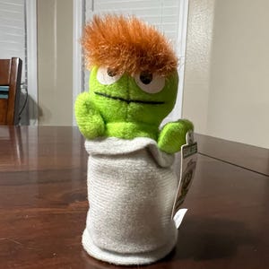 Puede incluir: Un peluche verde con pelo naranja, saliendo de un saco gris. El personaje tiene ojos grandes y una boca cosida. Una etiqueta con el logo de Sesame Street está adjunta.