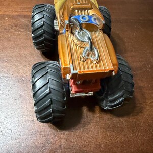 May include: A gold and black monster truck toy with large black tyres. The truck has a detailed design with a silver hook and blue accents. The toy is made of plastic and is on a wooden surface.