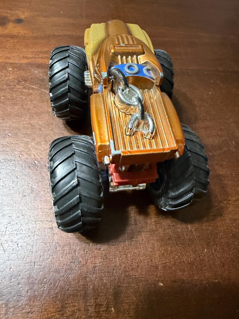 May include: A gold and black monster truck toy with large black tyres. The truck has a detailed design with a silver hook and blue accents. The toy is made of plastic and is on a wooden surface.
