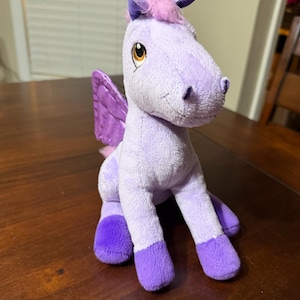 Puede incluir: Un peluche de pegaso en tonos de morado. El juguete tiene un cuerpo morado claro, pezuñas y alas moradas oscuras, y una melena y mechón rosas. Tiene ojos grandes y redondos y está sentado sobre una superficie de madera.