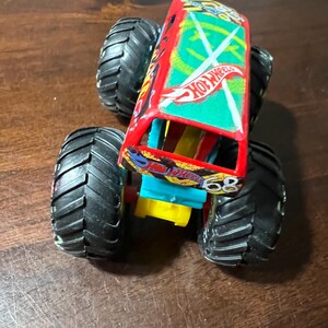 May include: A red and green Hot Wheels monster truck toy with large black tyres. The truck features the number 68 and the Hot Wheels logo. The body of the truck has a green and white design.