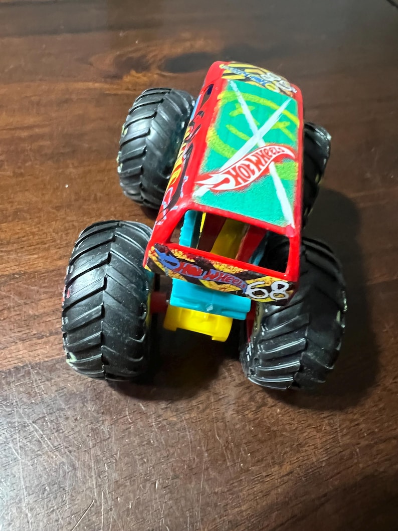 May include: A red and green Hot Wheels monster truck toy with large black tyres. The truck features the number 68 and the Hot Wheels logo. The body of the truck has a green and white design.