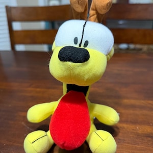 May include: A soft toy with a yellow body, a white head with brown ears, and a large red tongue. The toy has a black nose and eyes, and is on a wooden surface. The toy is a cartoon character.