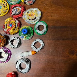 May include: A collection of colorful spinning tops, including red, yellow, green, and silver components. The tops have various designs and are arranged on a wooden surface. Some tops are partially assembled, showcasing their layered construction.