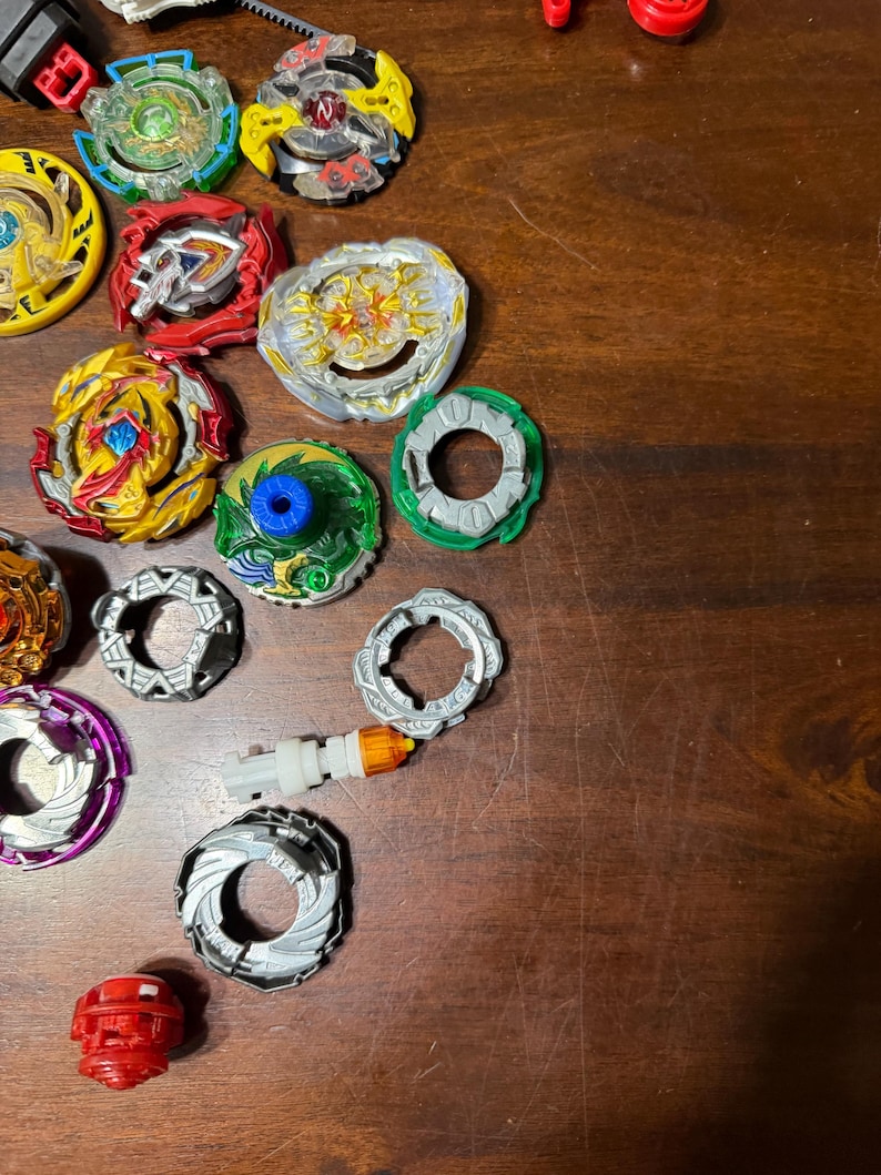 May include: A collection of colorful spinning tops, including red, yellow, green, and silver components. The tops have various designs and are arranged on a wooden surface. Some tops are partially assembled, showcasing their layered construction.