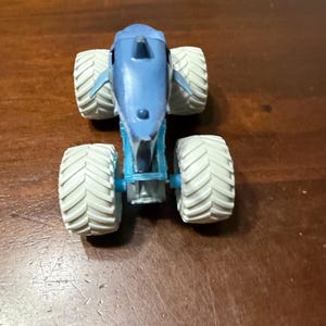 Puede incluir: Un juguete de cami&oacute;n monstruo azul con forma de tibur&oacute;n, con neum&aacute;ticos blancos grandes. El cuerpo del cami&oacute;n es azul claro. Los neum&aacute;ticos tienen un patr&oacute;n texturizado.
