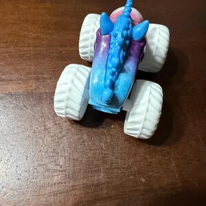 May include: A toy monster truck with a unicorn design. The truck has large white tyres and a body that fades from blue to purple. The unicorn features a blue mane and horn.