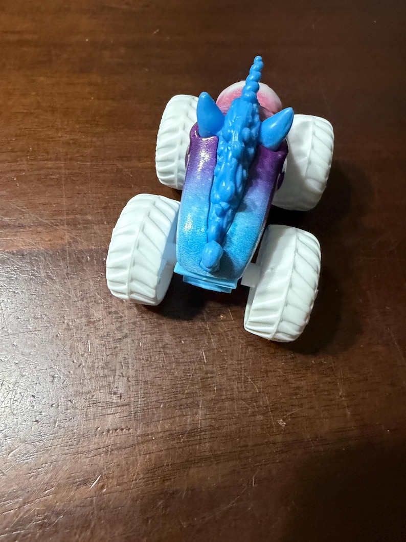 May include: A toy monster truck with a unicorn design. The truck has large white tyres and a body that fades from blue to purple. The unicorn features a blue mane and horn.