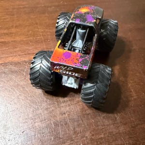 Pode incluir: Um brinquedo de monster truck com pneus pretos grandes. A carroceria do caminh&atilde;o &eacute; preta com desenhos de respingos de tinta laranja e roxa. As palavras "WILD SIDE" s&atilde;o impressas na lateral do caminh&atilde;o.