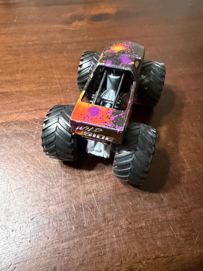 Pode incluir: Um brinquedo de monster truck com pneus pretos grandes. A carroceria do caminh&atilde;o &eacute; preta com desenhos de respingos de tinta laranja e roxa. As palavras "WILD SIDE" s&atilde;o impressas na lateral do caminh&atilde;o.