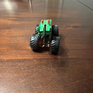May include: A green and orange monster truck toy with large black tires. The truck has a detailed design with a black chassis and orange accents. The toy is designed for play and collecting.