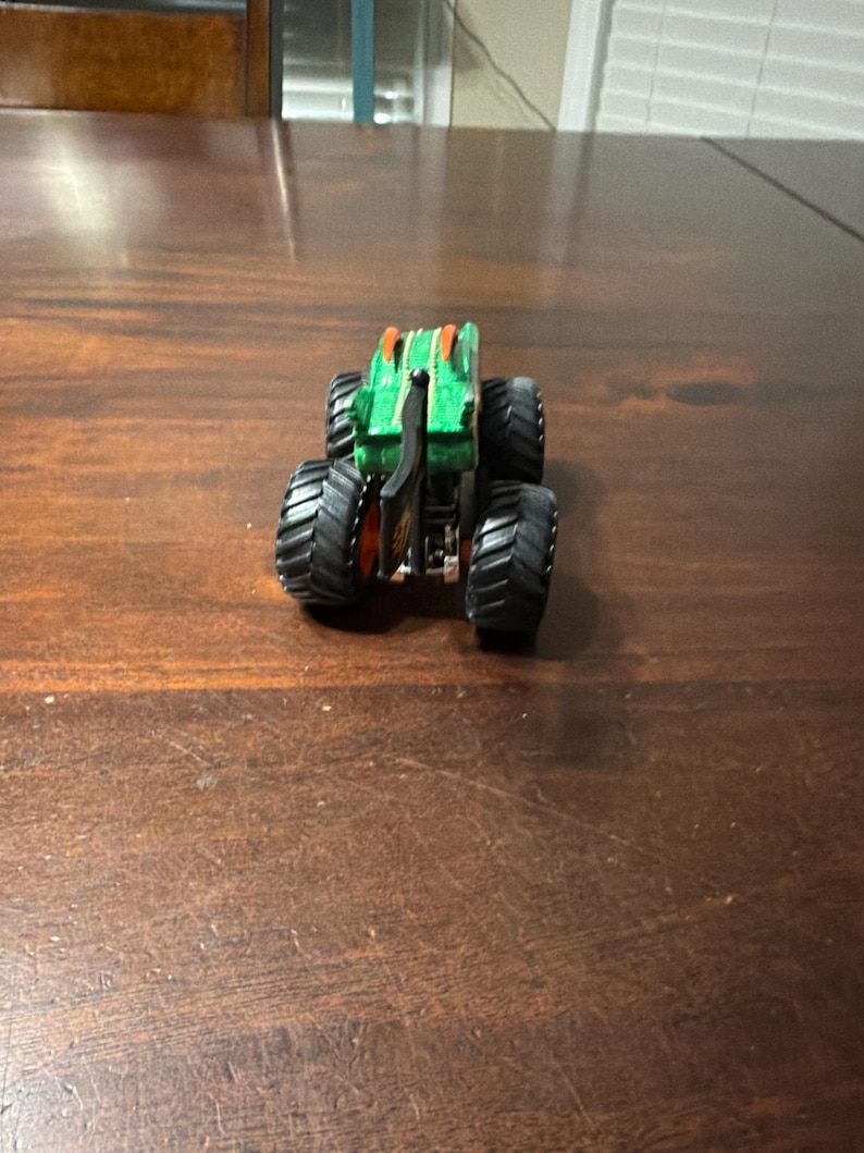 May include: A green and orange monster truck toy with large black tires. The truck has a detailed design with a black chassis and orange accents. The toy is designed for play and collecting.