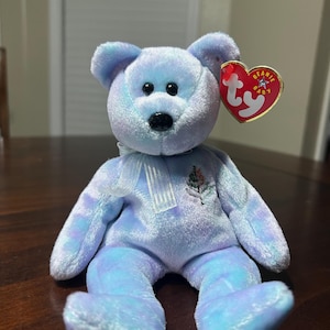 May include: A plush, light blue and purple teddy bear with a black nose and black eyes. The bear has a white ribbon around its neck and a red heart-shaped tag that reads "Beanie Baby".