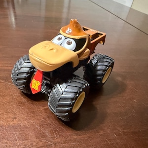 May include: A toy monster truck featuring a Donkey Kong character design. The truck has large black tyres with yellow rims, a tan face, and a brown hat. The red DK logo is visible on the front.